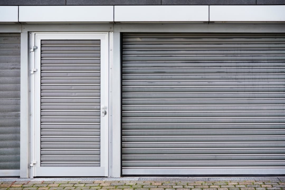 A modern storefront features a secure aluminum door alongside a closed metal roller shutter.