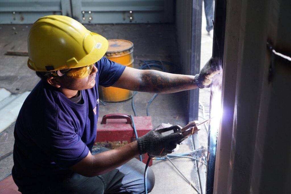 A worker wearing a yellow hard hat and safety glasses welds a metal shipping container frame.
